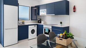 Cocina compacta con lavadora, microondas y horno, mesa de comedor con sillas y vista al mar a través de una ventana.
