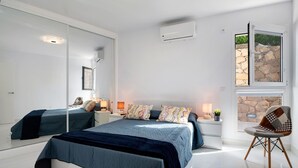 Quarto com cama, uma cadeira, janela com vista para uma parede de pedra, espelho e ar condicionado.