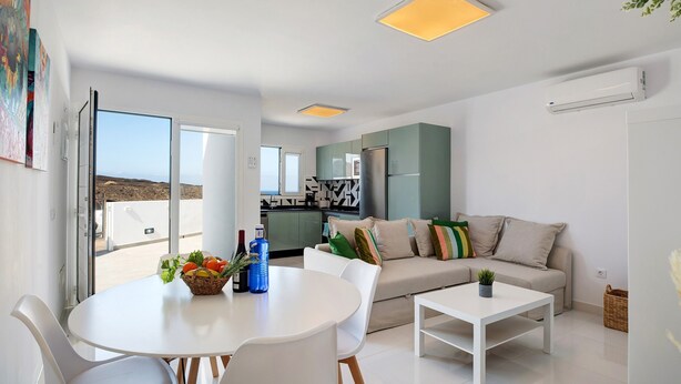 Uma sala de estar moderna com uma mesa de jantar, cadeiras brancas, um sofá e uma cozinha ao fundo.