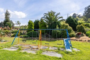 A playground with swings and a slide, surrounded by a garden with various plants and trees.