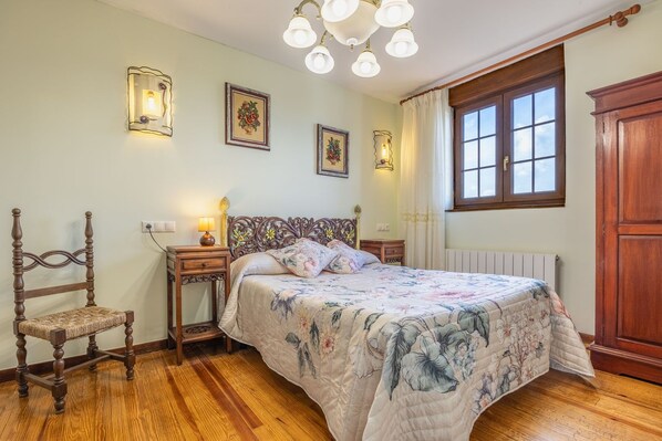 A bedroom with a wooden bed, a nightstand with a lamp, a chair, and a window with curtains.