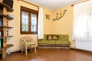 A room with a wooden floor, a sofa with patterned cushions, a wooden chair, a bookshelf, a window with curtains, and a wall with a floral painting.