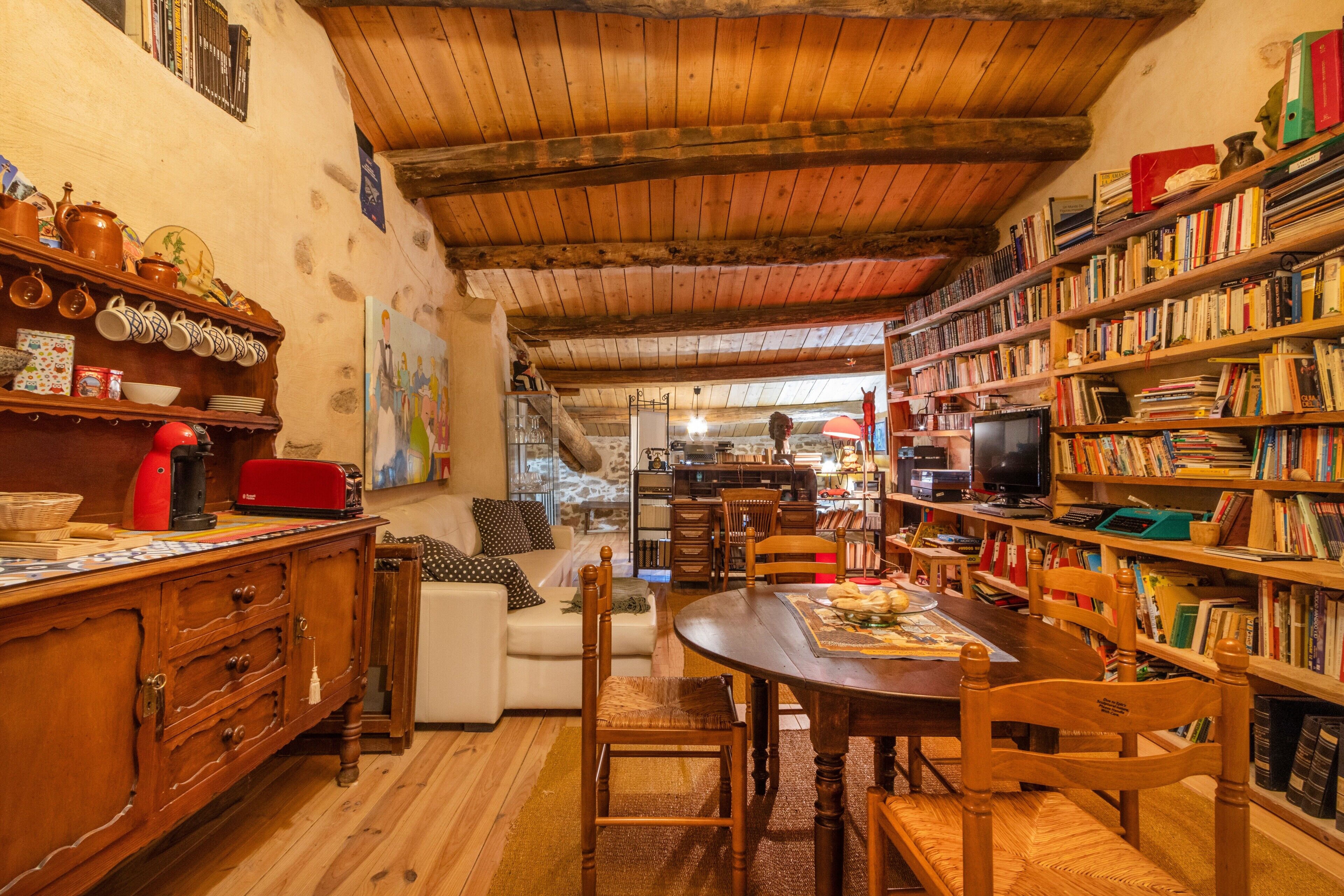 Een gezellige kamer met een houten plafond, een eettafel, stoelen, een boekenkast en een keukenhoek met een fornuis en diverse keukengerei.