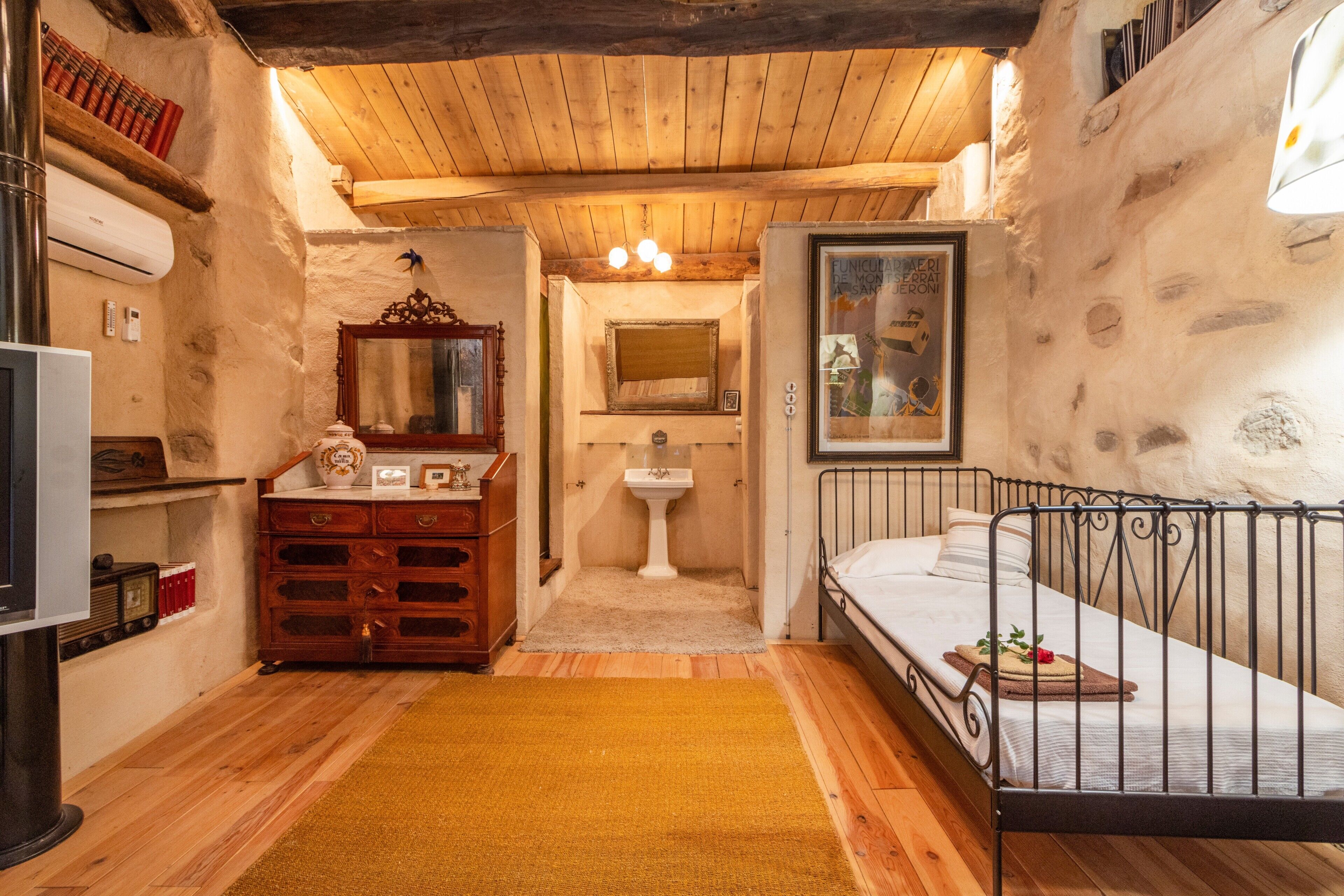 A rustic room with a wooden ceiling, a bed, a dresser, a mirror, a sink, and a television.