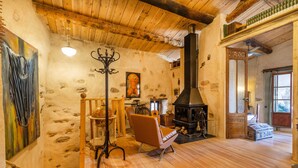 Una habitación rústica con pared de piedra, suelo de madera, chimenea, una silla y una escalera.