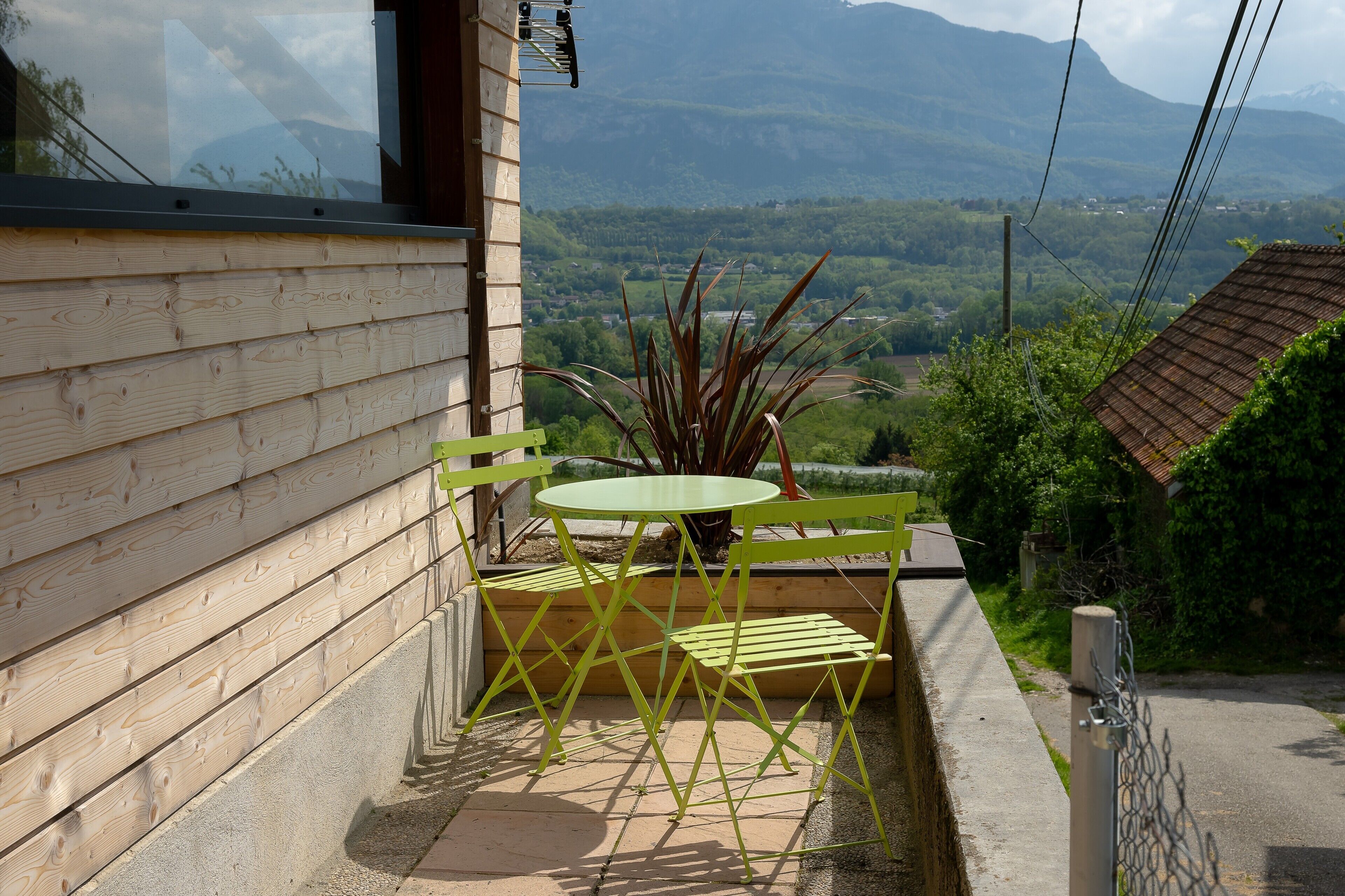 A wooden building with a balcony, a green metal table and chairs, and a view of a green landscape with mountains in the distance.