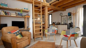 A cozy living room with a brown leather sofa, a wooden coffee table, and a colorful chair. A wooden ladder is leaning against the wall, and there are shelves with books and decorative items. - Holiday Home 'Le Balcon Des Bauges' with Mountain View, Shared Pool and Wi-Fi (LA MOTTE-SERVOLEX)