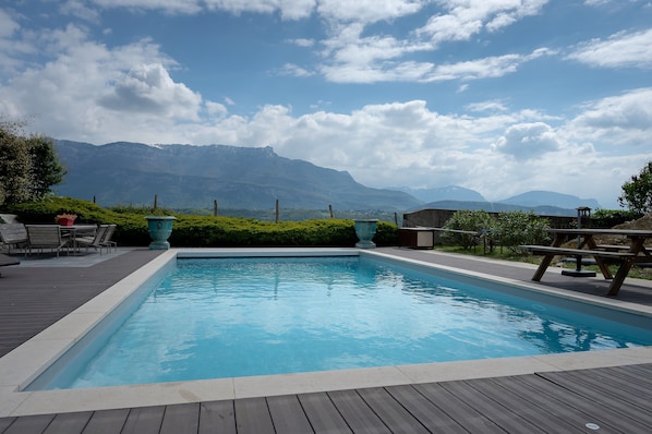 Una piscina con cubierta de madera, mobiliario exterior y vistas a montañas.