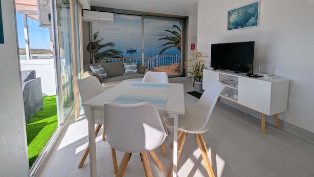 Una sala de estar moderna con comedor, televisor y un amplio ventanal con vista al mar.