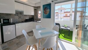 Cocina moderna con armarios blancos, microondas y cafetera. Cuenta con un comedor con mesa y sillas, además de un amplio ventanal con vistas a un césped verde y edificios.