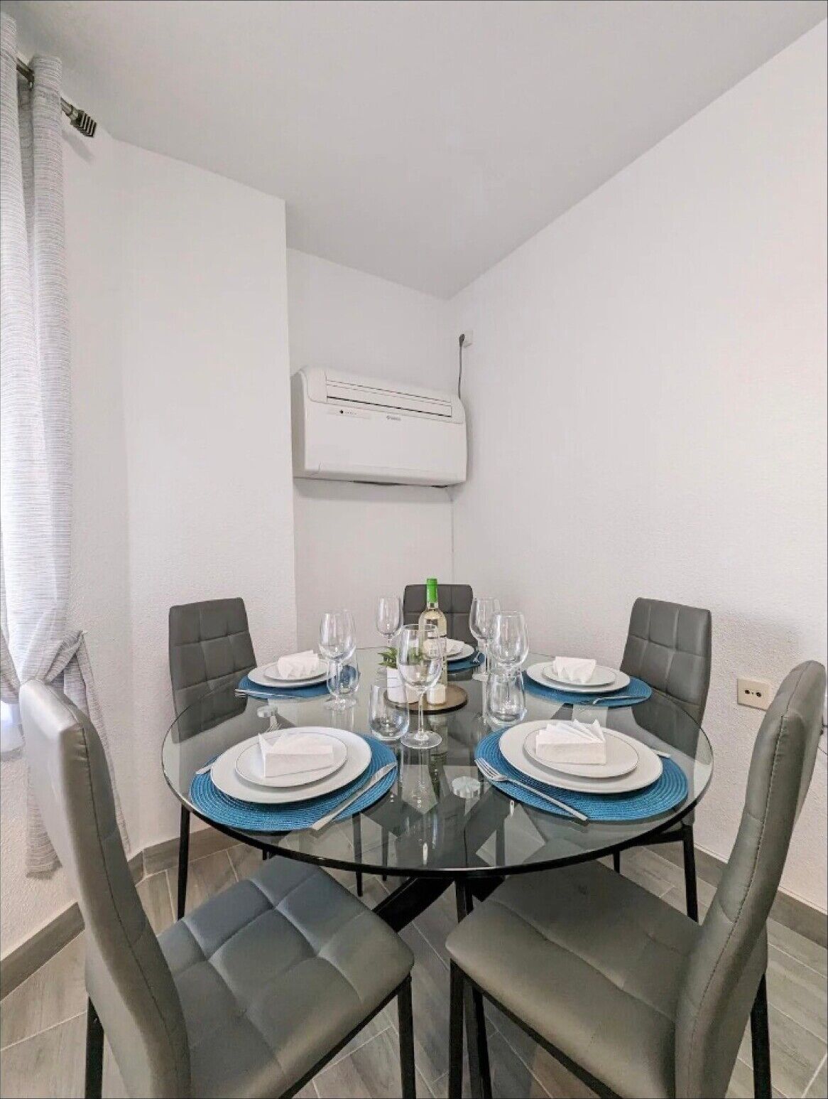 Un comedor con una mesa de vidrio, cuatro sillas grises y un equipo de aire acondicionado blanco en la pared.