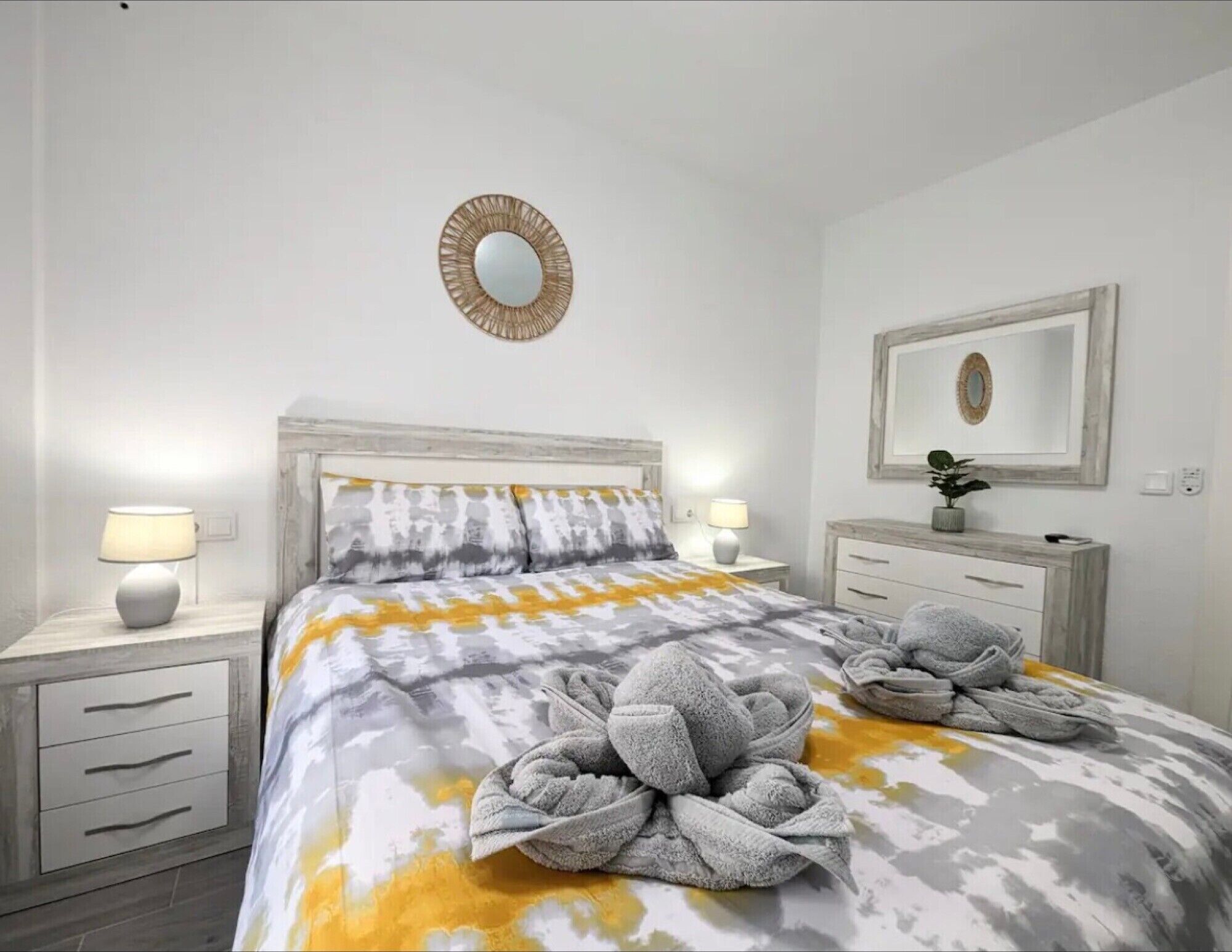 Una cama bien hecha con una colcha estampada, dos lámparas de noche, una mesita de noche con cajón, un espejo en la pared y una pequeña planta en maceta.
