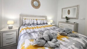 Una cama bien hecha con una colcha estampada, dos lámparas de noche, una mesita de noche con cajón, un espejo en la pared y una pequeña planta en maceta.