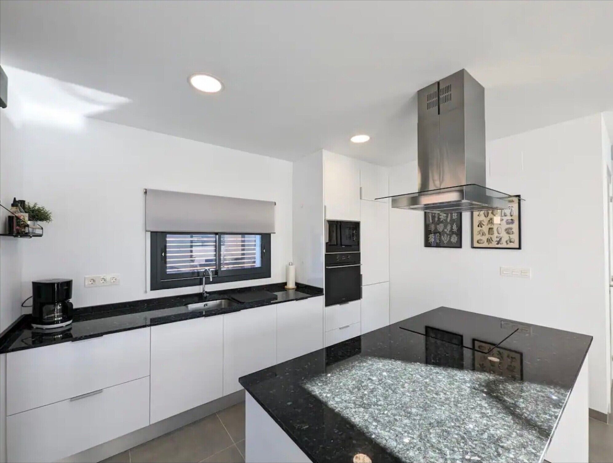 Una cocina moderna con armarios blancos, encimera negra, ventana con persianas y una campana extractora de acero inoxidable.