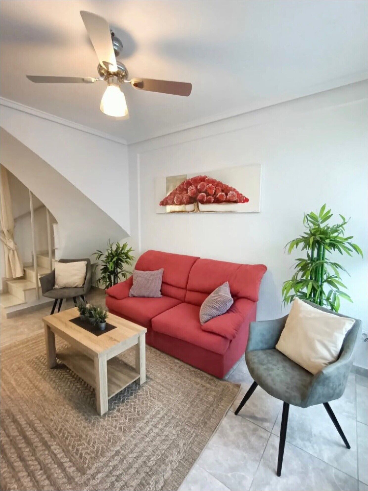 A living room with a red sofa, a wooden coffee table, a grey armchair, a ceiling fan, a staircase, and a wall-mounted artwork.