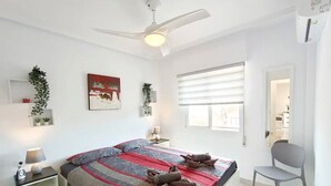 Un dormitorio con cama, mesitas de noche, ventilador de techo, ventana con persianas, una silla y una planta pequeña.