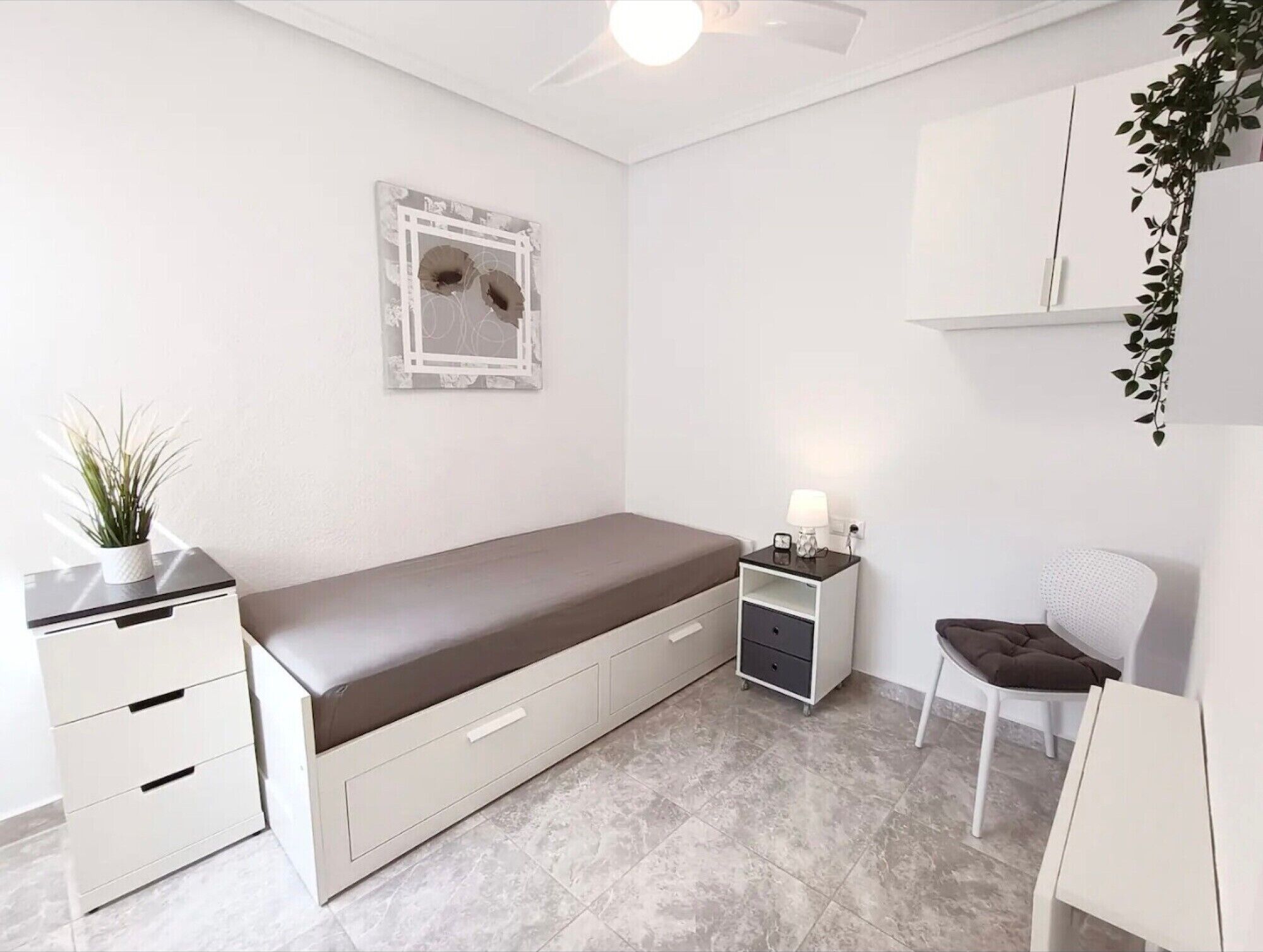 Une pièce minimaliste comprenant un lit, un petit bureau, une chaise, une table de chevet, une étagère fixée au mur et une plante en pot.