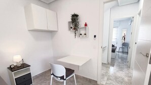 Una habitación pequeña y blanca con un escritorio, una silla y una mesita de noche con lámpara. Hay una puerta que da a otra habitación.