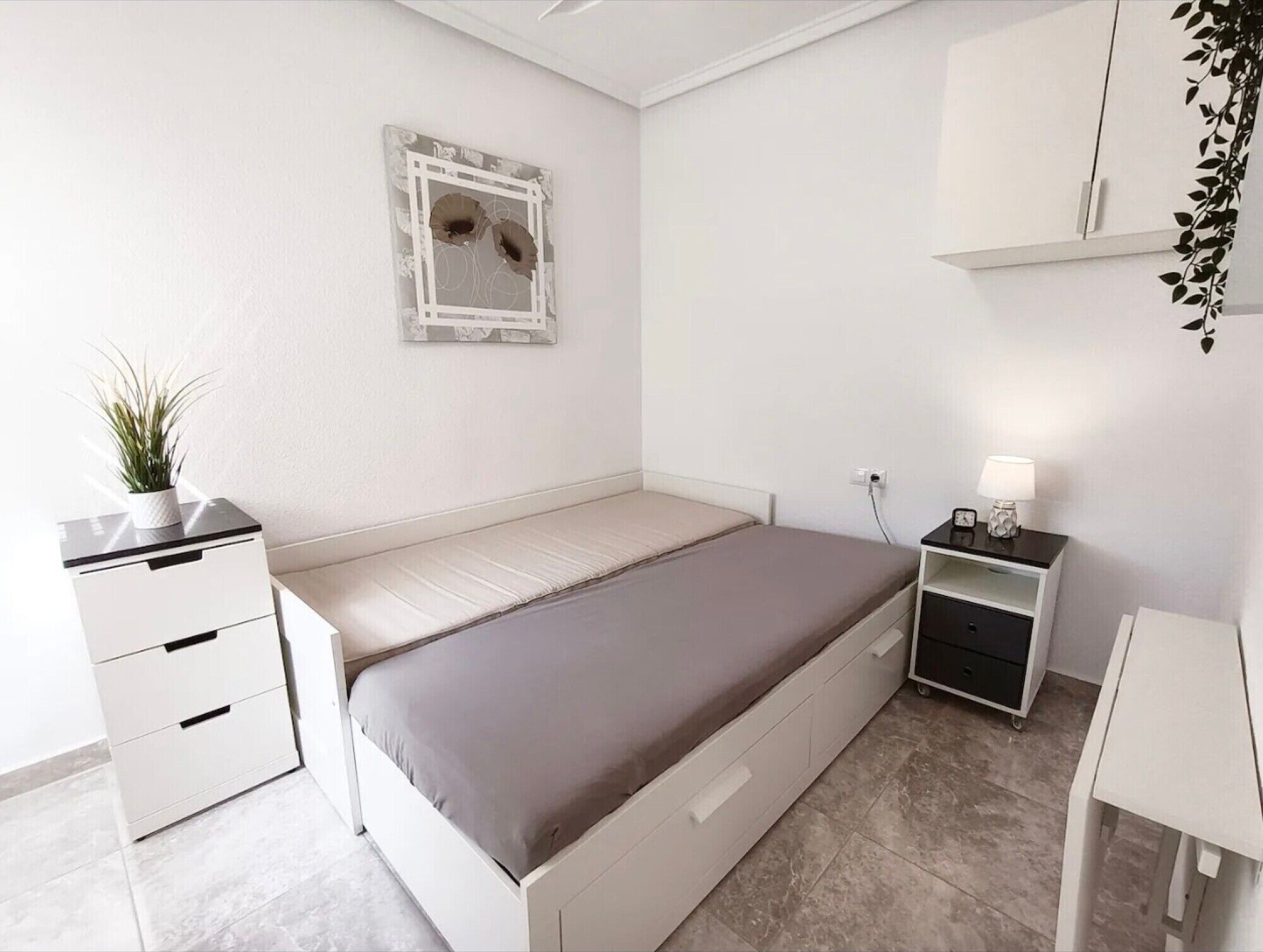 A bedroom with a bed, bedside tables, a nightstand, a wall-mounted mirror, and a potted plant.