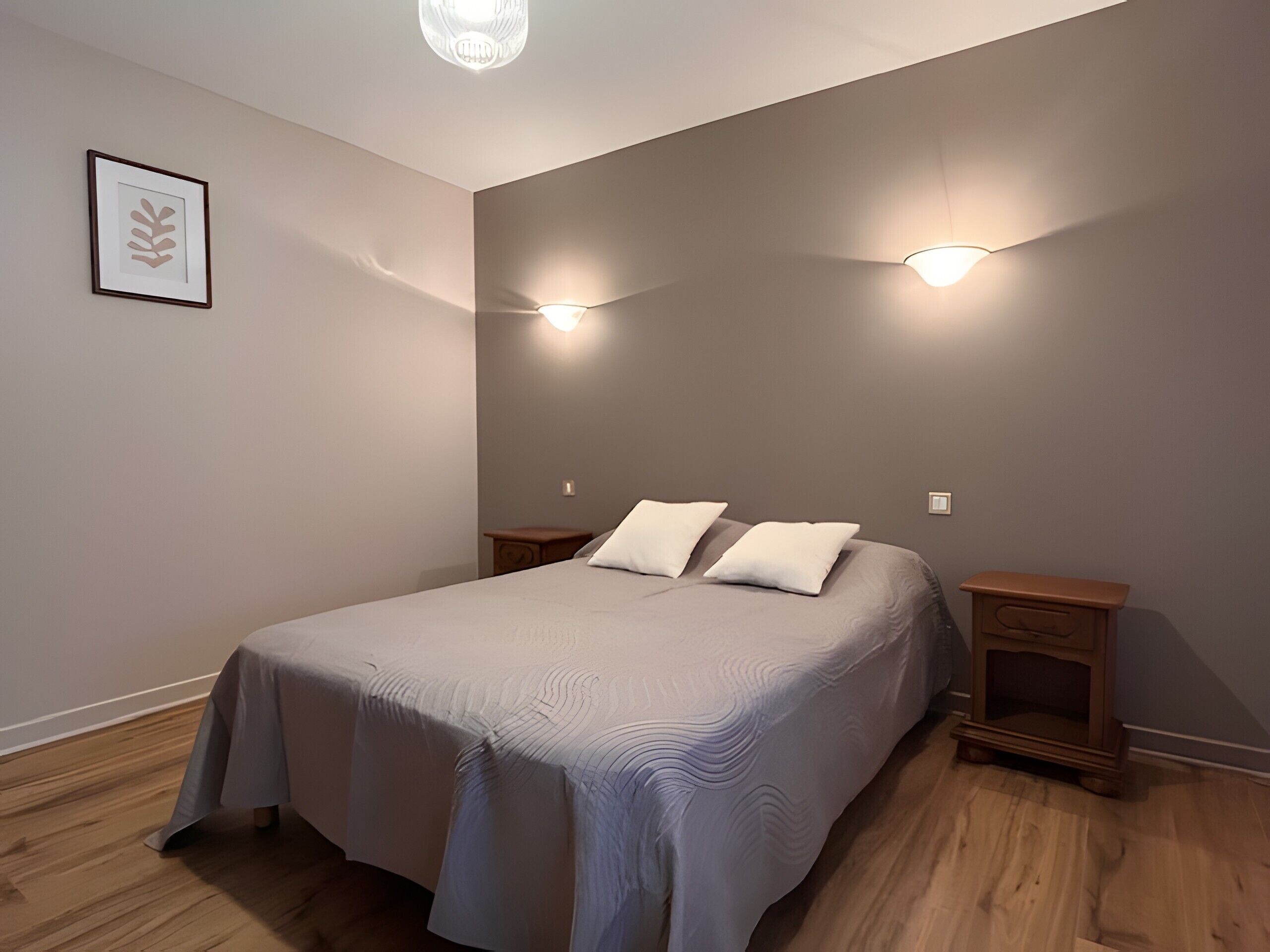 A bedroom with a bed, two bedside tables, a nightstand, a wall-mounted light, and a framed picture.