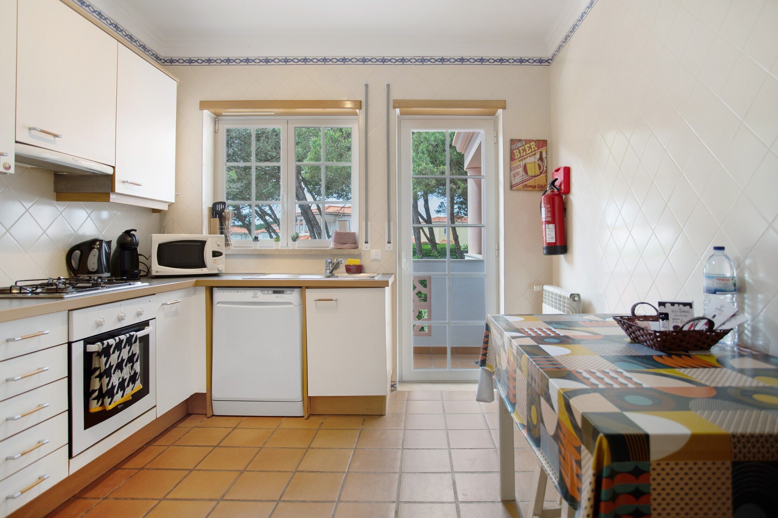 Ett kompakt kök med mikrovågsugn, spis och diskmaskin, ett matbord med en färgglad duk och ett fönster med utsikt över en trädgård.
