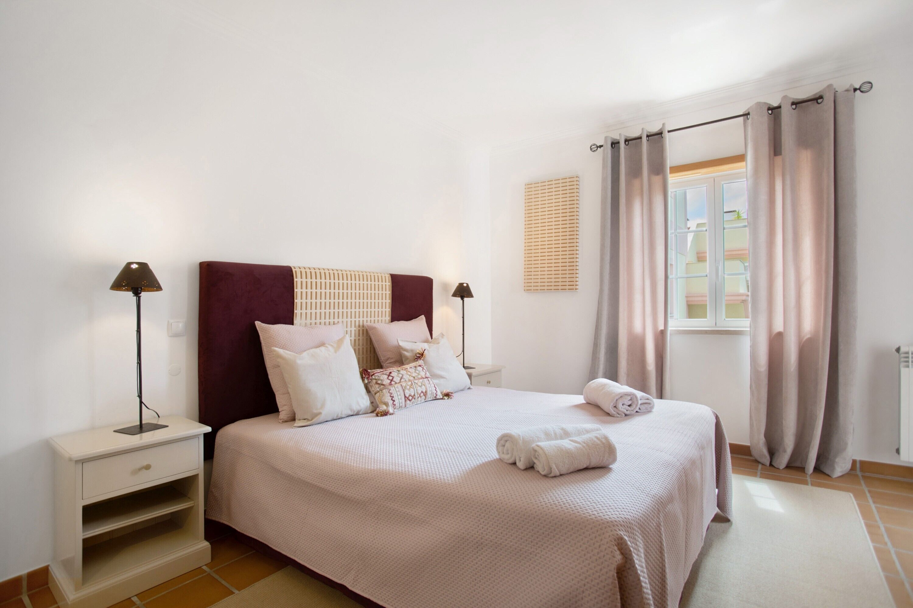 Une chambre à coucher bien rangée, avec un lit, des tables de chevet, une fenêtre avec des rideaux et un luminaire fixé au mur.
