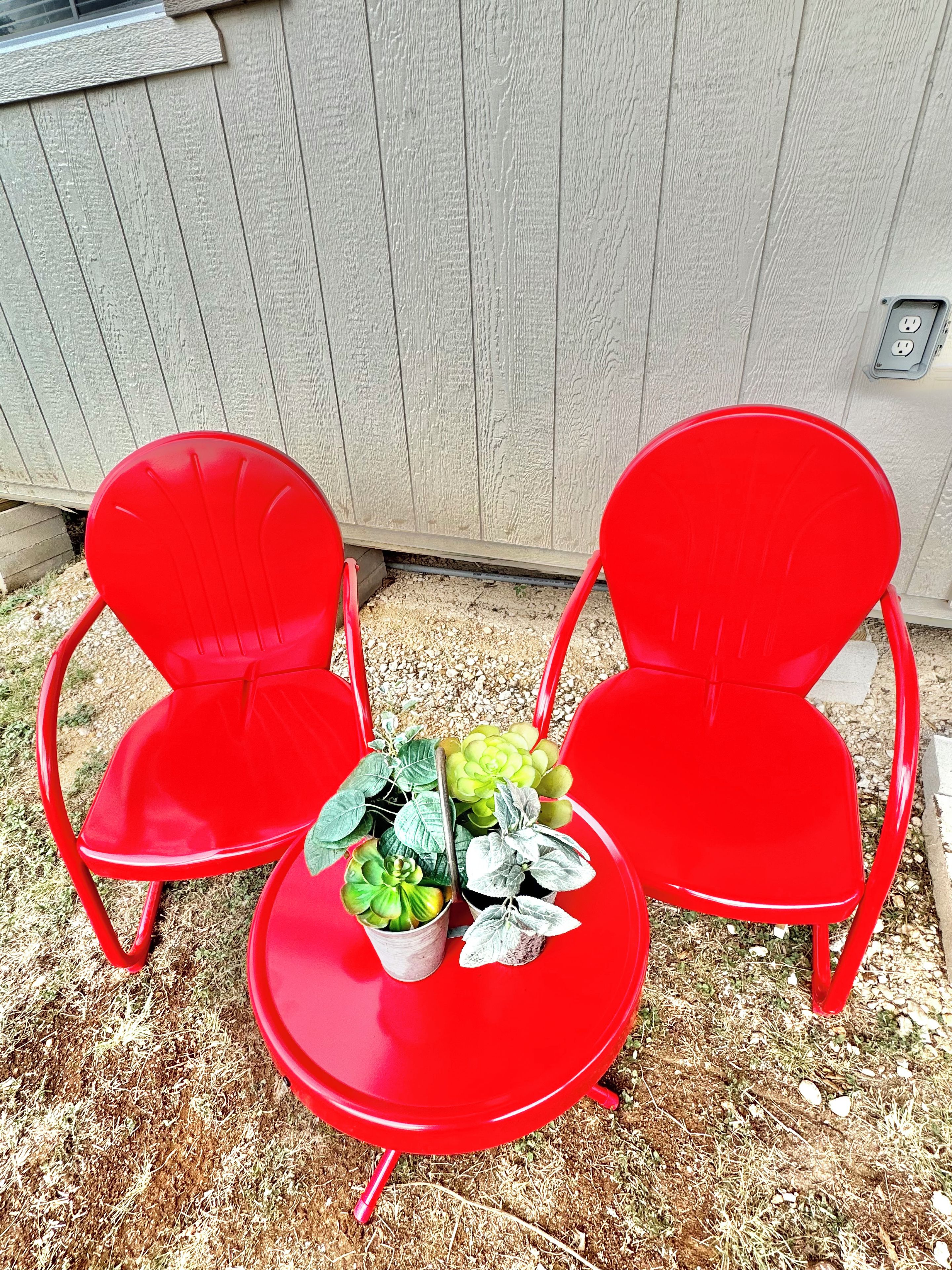 Two red chairs and a small table with potted plants in front of a wooden fence.