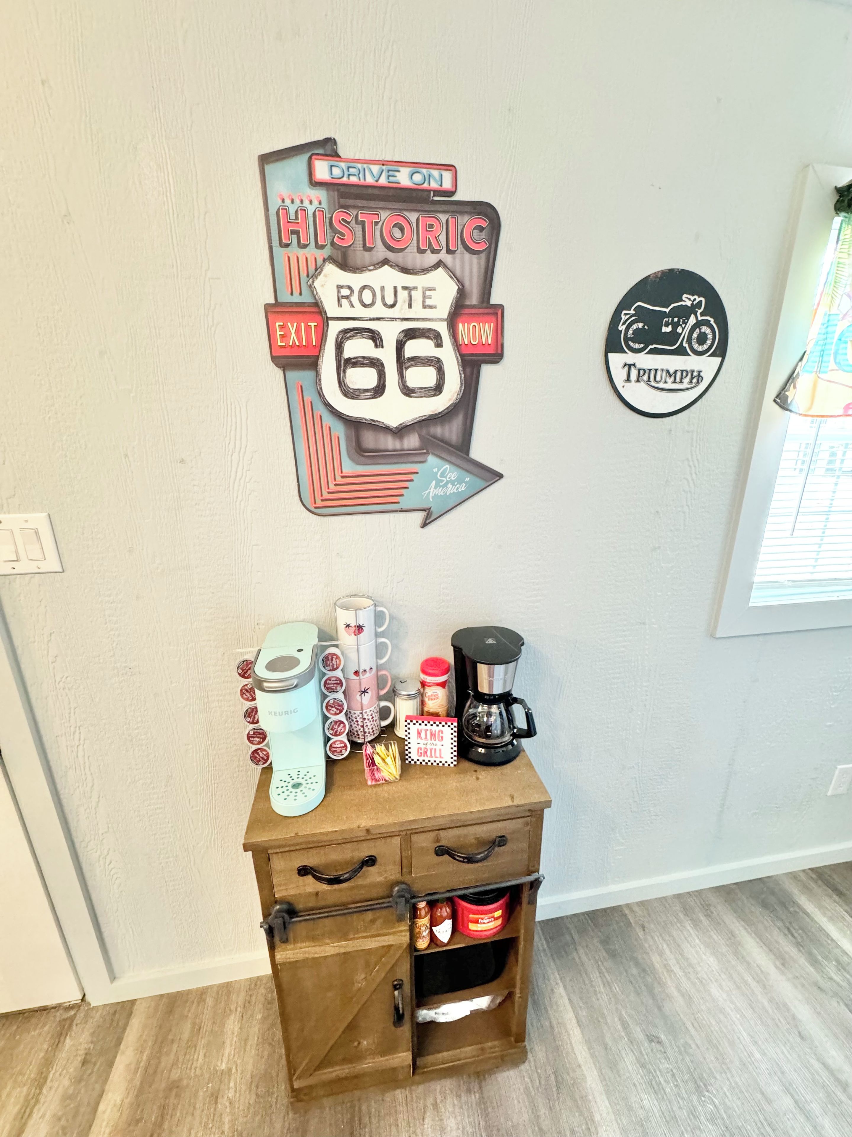 Un mur orné d’un panneau indiquant la Route 66 et d’un panneau représentant une moto Triumph. En dessous, sur un meuble en bois, on aperçoit une machine à café et des dosettes Keurig.