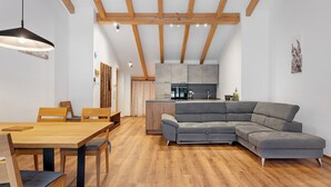 Un intérieur moderne comprenant un coin repas, un canapé d’angle gris, des poutres en bois et une cuisine.