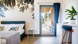 A compact hotel room with a bed, a small stool, a desk with a chair, a potted plant, and a view of the outside. - Casale Lorenzo - Apartment 1 with Private Terrace, Shared Garden and Air Conditioning (Orbetello)