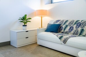A white sofa with a leaf-patterned throw, a bedside table with a lamp, and a potted plant.
