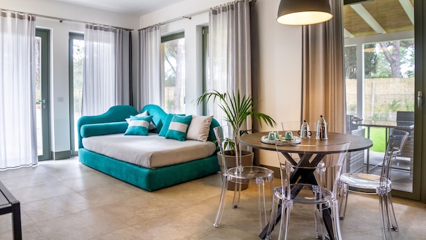 A modern living room with a teal sofa, a coffee table, clear chairs, and a large window with curtains. - Casale Lorenzo - Apartment 2 with Private Terrace, Shared Garden and Air Conditioning (Orbetello)