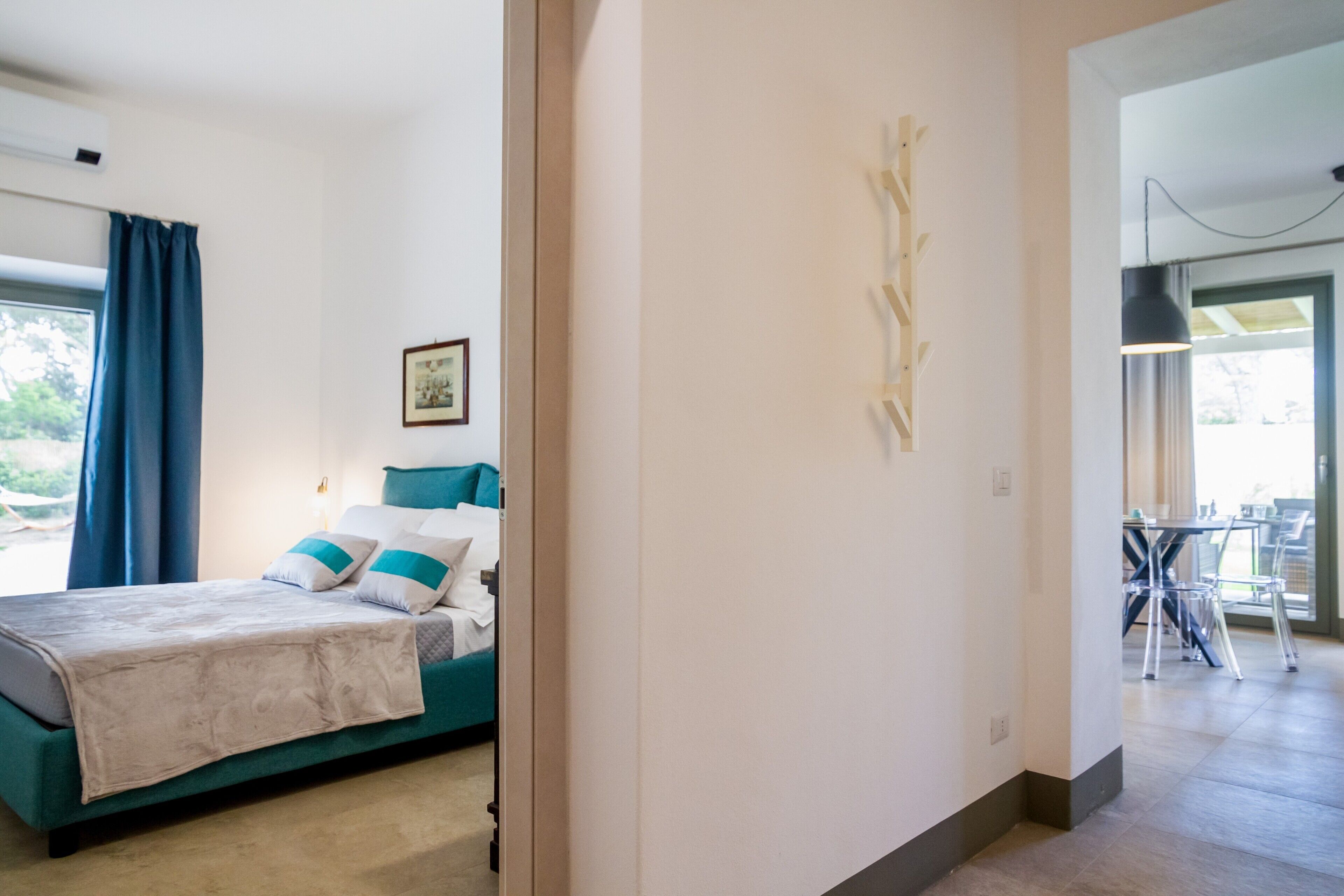 Une chambre à coucher avec un lit, une fenêtre avec des rideaux, un tableau encadré au mur et un couloir menant à une salle à manger.