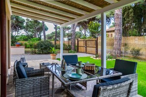 An outdoor patio with wicker furniture, a glass table, and a view of a garden and trees.