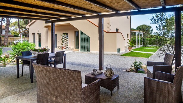 Un patio exterior con mobiliario de mimbre, una zona cubierta con techo de madera y vista a un edificio con una puerta verde.