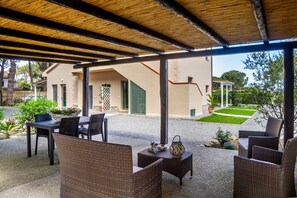 An outdoor patio with wicker furniture, a covered area with a wooden ceiling, and a view of a building with a green gate.
