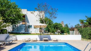 Una piscina con sillas de descanso, un edificio blanco con balcón y un árbol con flores moradas.