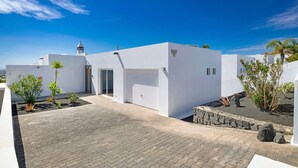 Un bâtiment moderne de couleur blanche, doté d’un garage, d’une petite tour et d’une allée pavée agrémentée de plantes.