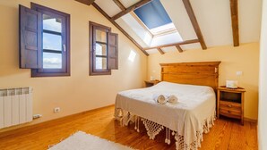 Un dormitorio con tragaluces, vigas de madera, una cama con colcha blanca, una mesita de noche y una mesilla.