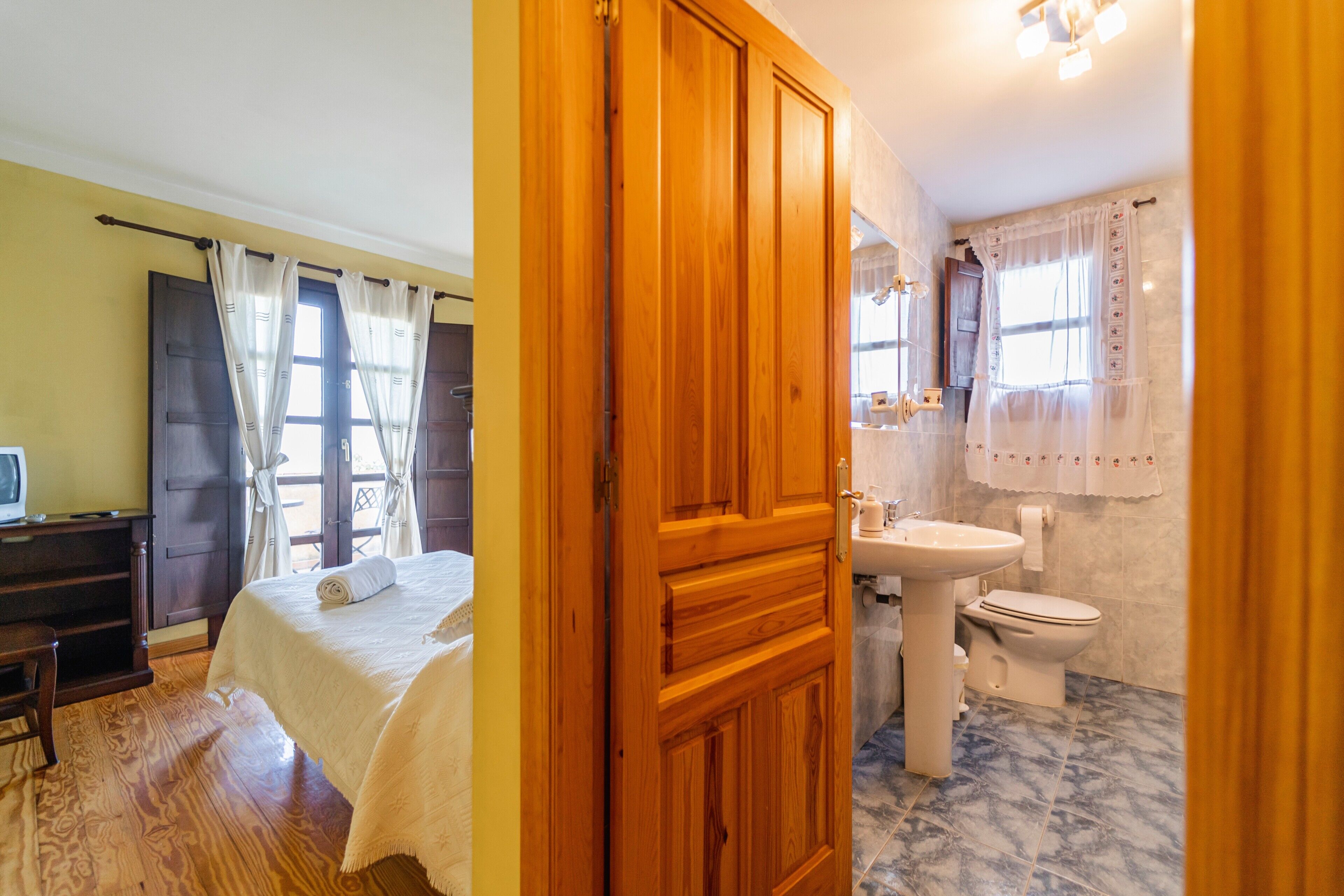 A bedroom with a bed, a wooden door, a desk, and a window with curtains.