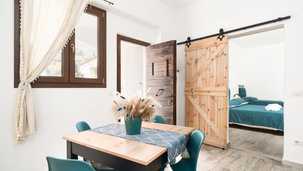 A room with a wooden table, teal chairs, a wooden door, and a bed with a green bedspread. - Apartment 'S' Istancu Vetzu' with Mountain View, Wi-Fi and Air Conditioning (Irgoli)