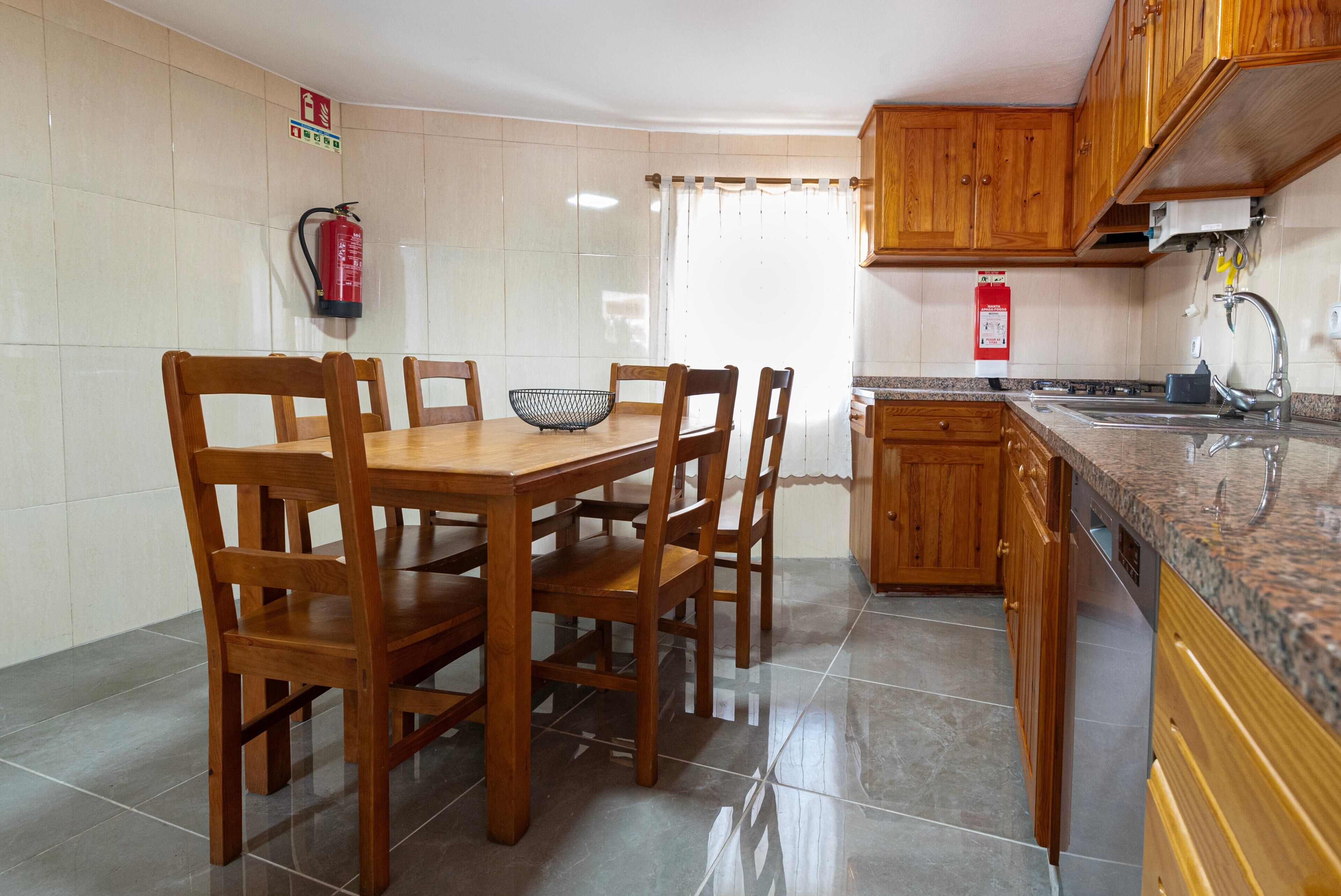 Uma cozinha com armários de madeira, chão de azulejos, mesa de jantar com cadeiras, extintor de incêndio e janela com cortinas.