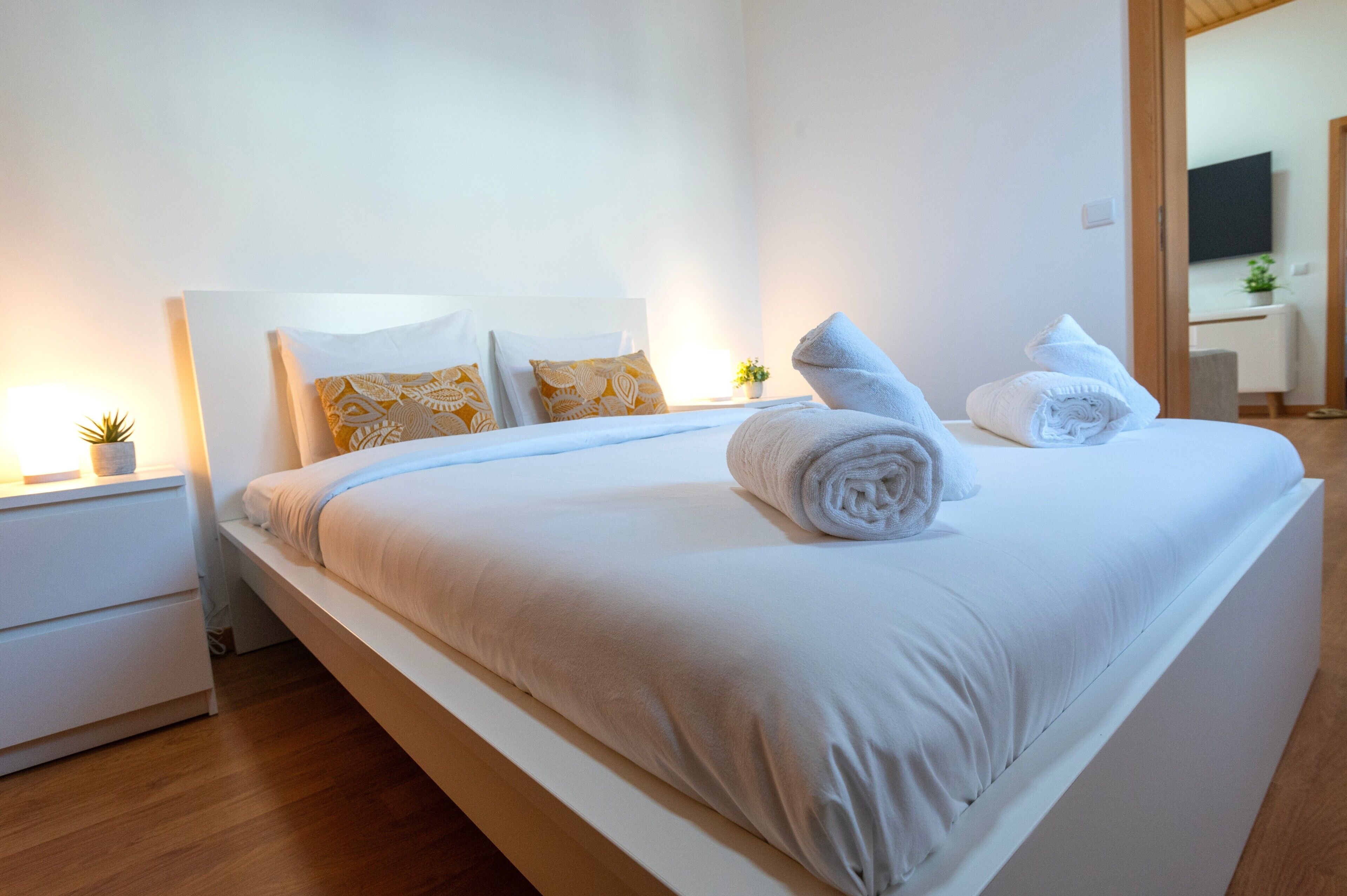 A neatly made bed with white linens and rolled towels, a bedside table with a lamp, and a small potted plant.