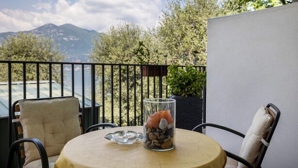 A round table with a glass vase containing rocks and a decorative item, a chair with a cushion, and a view of mountains and trees through a balcony railing. - Studio Apartment 'Tiny' with Shared Terrace and Wi-Fi (Brenzone)