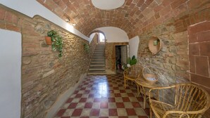 A narrow hallway with a checkered floor, stone walls, and a staircase leading to an upper level. There are wicker chairs and baskets along the right side.