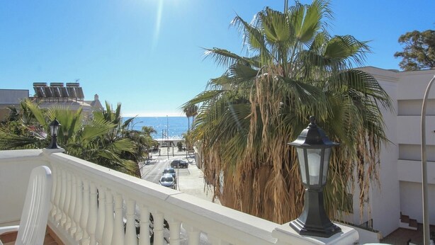 Een balkon met een hekwerk, palmbomen, een straat met auto's en uitzicht op zee.