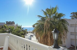Un balcon avec une balustrade, des palmiers, une rue avec des voitures et une vue sur la mer.