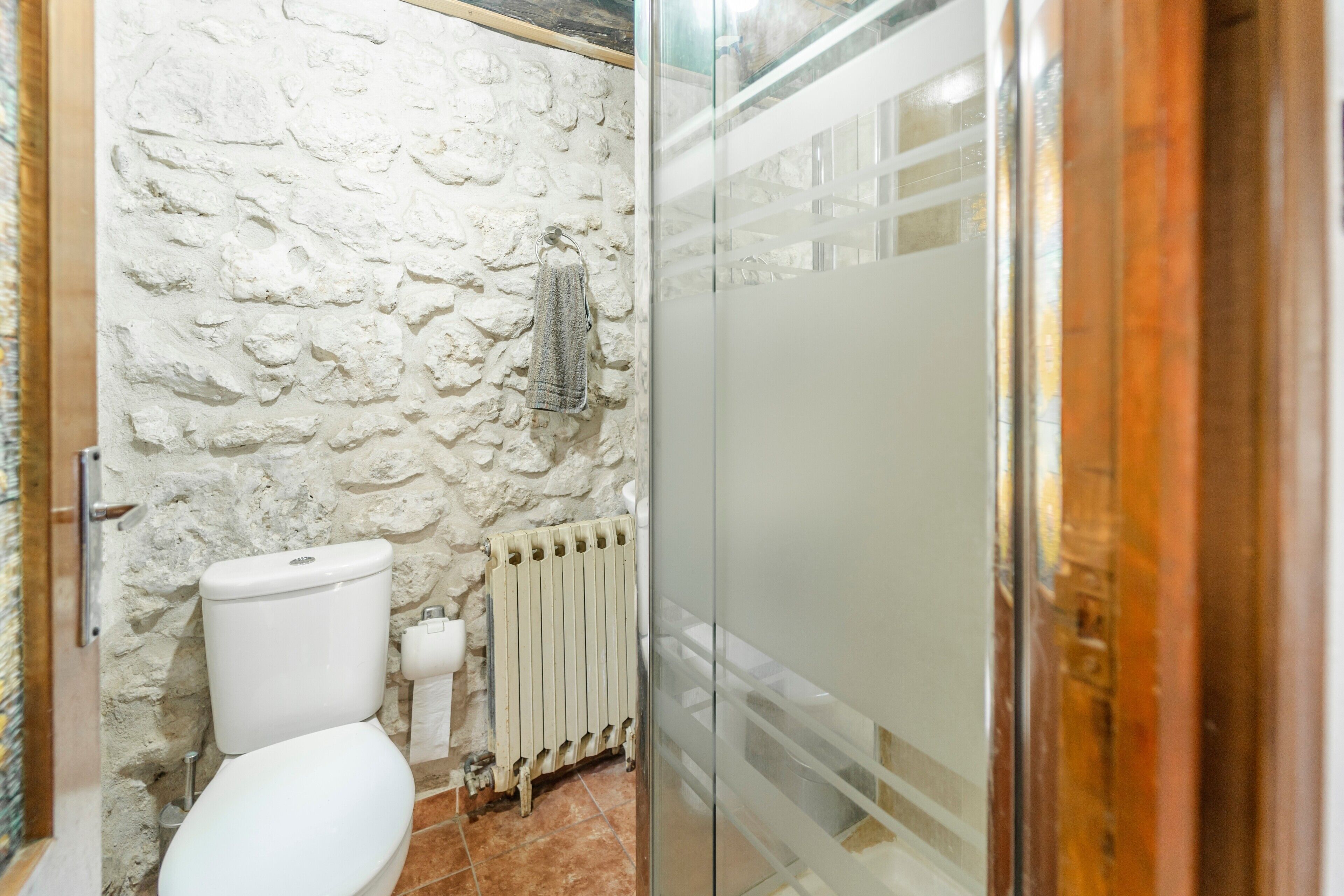 A bathroom with a stone wall, a toilet, a radiator, and a glass shower enclosure.