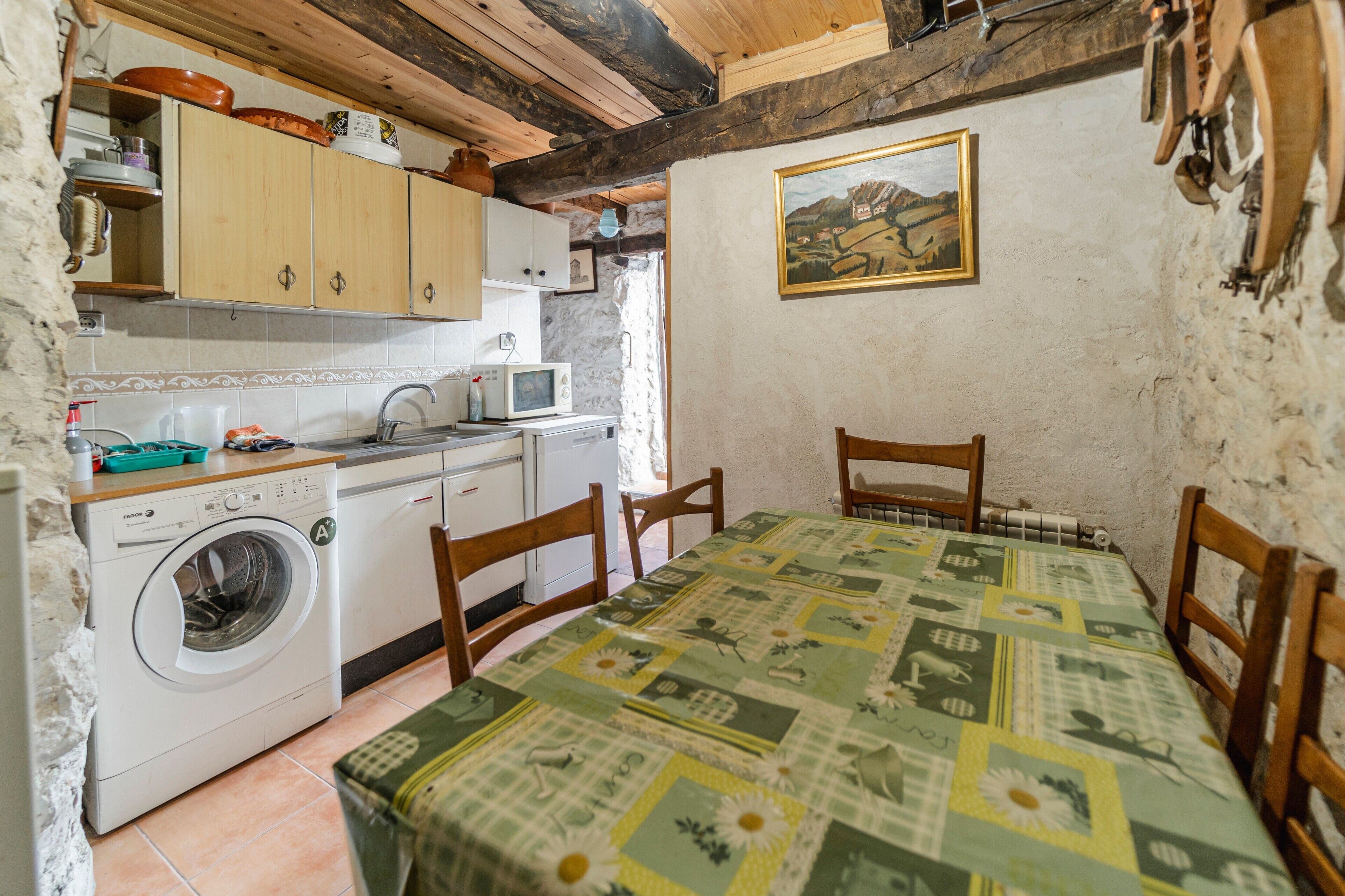 A compact kitchen with a washing machine, wooden cabinets, a microwave, and a table with a patterned tablecloth.