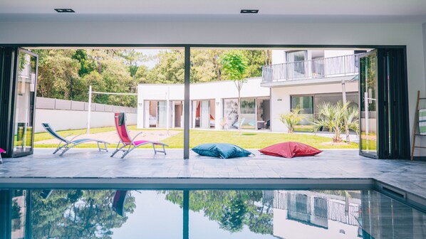 A modern indoor pool area with a glass enclosure, lounge chairs, and a view of a residential building.